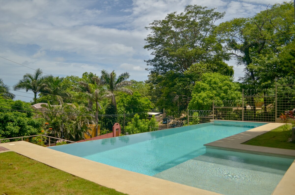 An infinity pool is situated within a lush tropical landscape, surrounded by greenery and mature trees. The clear blue water reflects the sky, creating a serene atmosphere. Adjacent to the pool, a well-maintained lawn enhances the natural beauty of the setting.