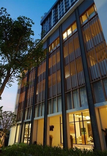 The exterior of the modern building is showcased, with its floor-to-ceiling windows framed by wooden slats. A well-maintained garden with greenery borders the lower level, complementing the sleek design. The soft glow from the interior lights adds to the inviting atmosphere of the structure.