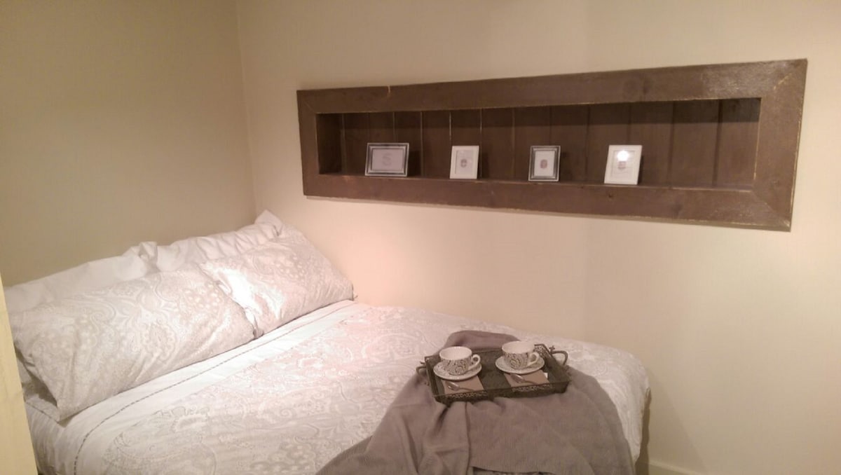 A cozy sleeping area features a spacious bed with light-colored bedding. A wooden shelf above the bed holds framed pictures, while a tray at the foot of the bed is set with two cups and a small plate. Soft lighting enhances the warm atmosphere.