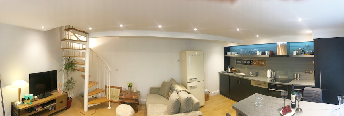 An open-plan living space is showcased, featuring a light wooden floor and well-lit areas. A comfortable sofa is positioned near a coffee table, with a television and a vintage-style refrigerator visible. A staircase leads to the upper level, adding vertical interest to the room.