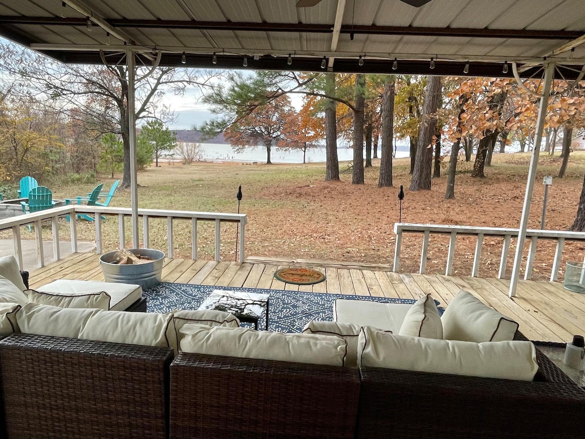 A large back deck features plush seating arranged around a central coffee table. A fire pit and decorative items are placed nearby. The view showcases a serene expanse of lake and trees, enhancing the outdoor living experience.