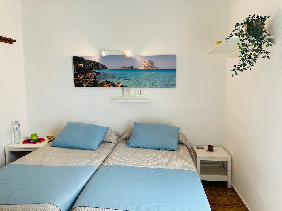A compact sleeping area is shown, featuring two separate beds adorned with blue bedding. A scenic wall art piece displays a coastal view, while a small bedside table holds a water bottle and a decorative item. Soft natural light fills the space.