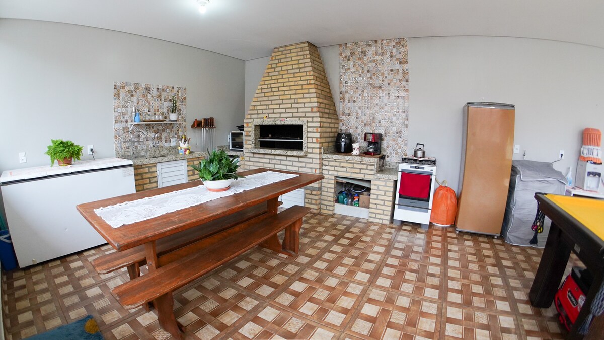 The kitchen area features a large wooden dining table with ample seating. A brick fireplace is positioned against a decorative tiled wall. Basic kitchen appliances are visible, including a refrigerator, and there is a spacious countertop for food preparation.