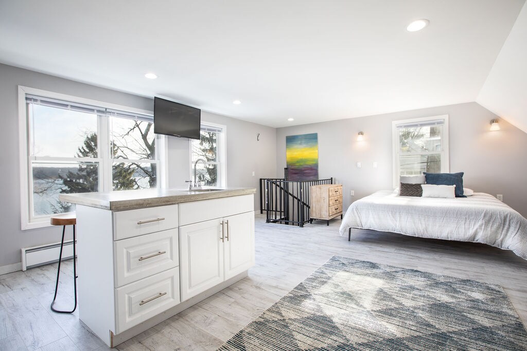A studio space is displayed with natural light flooding through large windows, revealing views of Irondequoit Bay. A white kitchen island with a countertop is positioned near the center, while a comfortably made bed with neutral linens is situated against the wall, and a colorful artwork is mounted above.