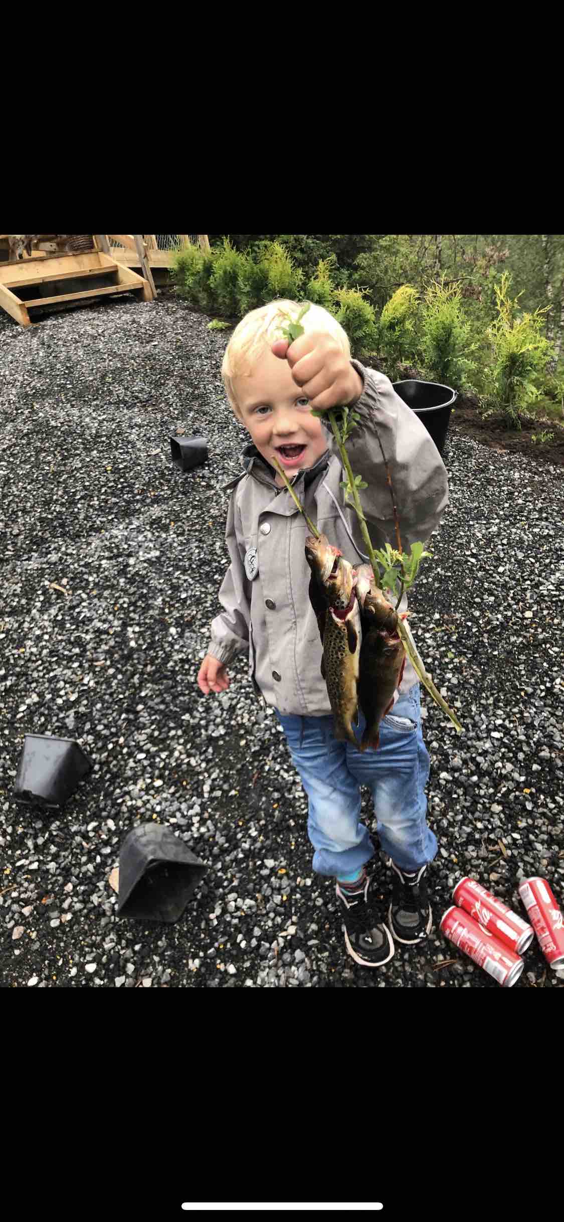 A child stands outdoors, holding several fish by their tails, with greenery in the background. The setting includes a gravel surface scattered with empty cans and potted plants. The child's clothing consists of a light jacket and jeans.