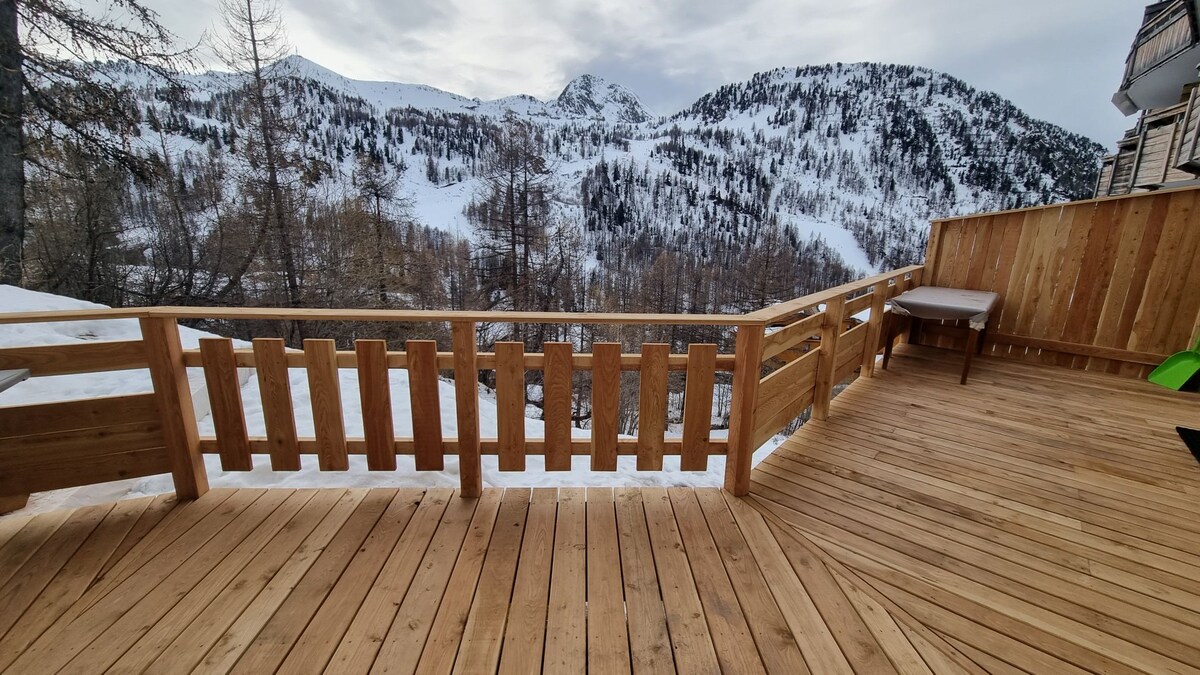 A spacious wooden terrace is presented, showcasing a scenic view of the mountains blanketed in snow. The open space features wooden railings and a small table is positioned on the terrace, inviting relaxation while enjoying the surrounding natural beauty.