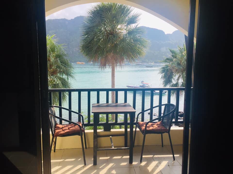 A viewpoint features a balcony with two metal chairs and a small table. Lush palm trees frame the scene, overlooking a clear body of water where a boat is visible in the distance against a backdrop of mountains.