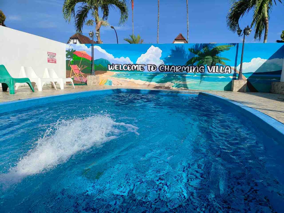 A large private pool is framed by a vibrant mural depicting a beach scene. White lounge chairs are positioned nearby, inviting relaxation. Palm trees line the area, contributing to the overall tropical ambiance surrounding the villa.