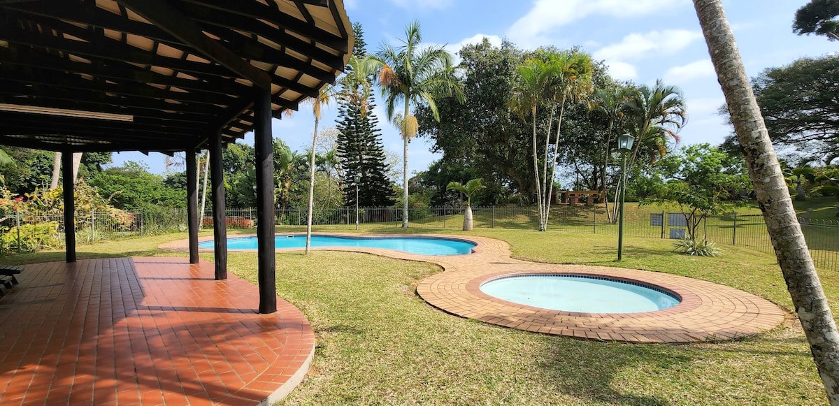 A spacious outdoor area is depicted, featuring two swimming pools surrounded by greenery. The larger pool is accompanied by a smaller circular spa. Palm trees and well-maintained grass create a serene environment, with gentle sunlight filtering through the trees.