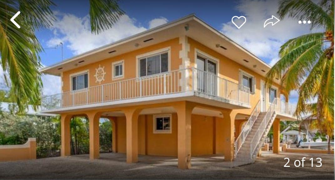 A two-story house is presented with a bright orange exterior. The structure features a balcony and large windows, providing ample natural light. Palm trees frame the property, enhancing the tropical environment. A gravel area surrounds the house, contributing to the overall spacious feel.