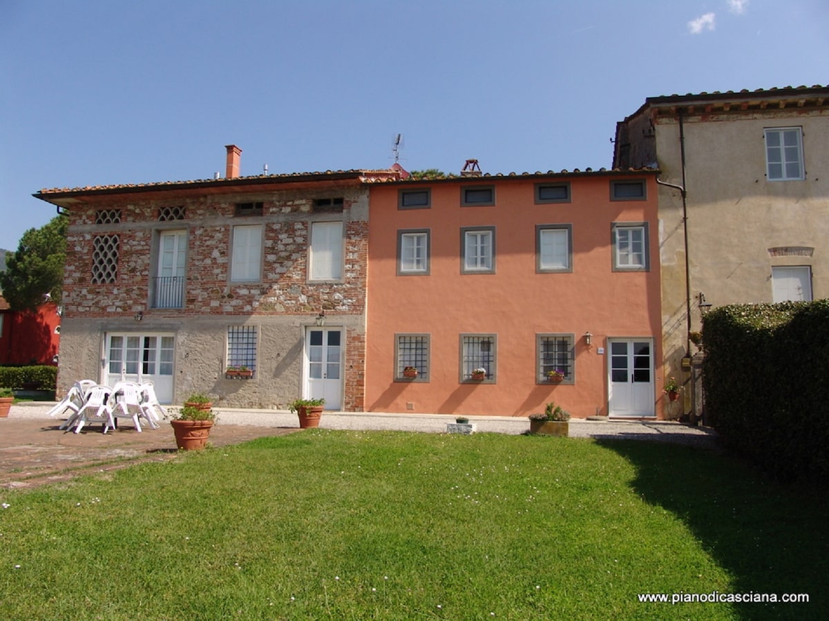 A two-story rustic building features a combination of stone and plaster in warm colors. Window boxes with flowers add charm to the facade. A grassy area with white chairs is visible in the foreground, complementing the tranquil outdoor setting.
