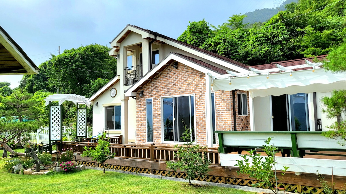 The two-story villa features a brick facade with large windows for natural light. A spacious deck wraps around, complemented by a gazebo and well-maintained garden. Lush greenery surrounds the property, adding to the tranquil environment.