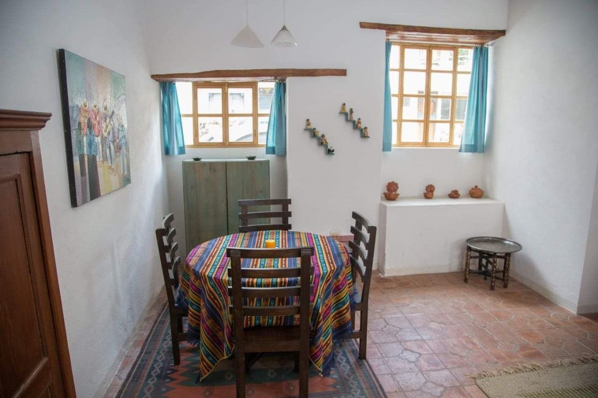 A dining area is presented with a colorful tablecloth draping over a round wooden table surrounded by four chairs. Light filters through two windows adorned with sheer blue curtains, highlighting a wall featuring decorative pieces and a small shelf displaying pottery.