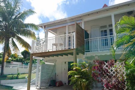 The exterior of a two-story apartment is visible, featuring a spacious balcony with a small table and chairs. Palm trees and vibrant tropical plants surround the area, while bright sunlight enhances the welcoming atmosphere. The white building displays traditional architectural details.