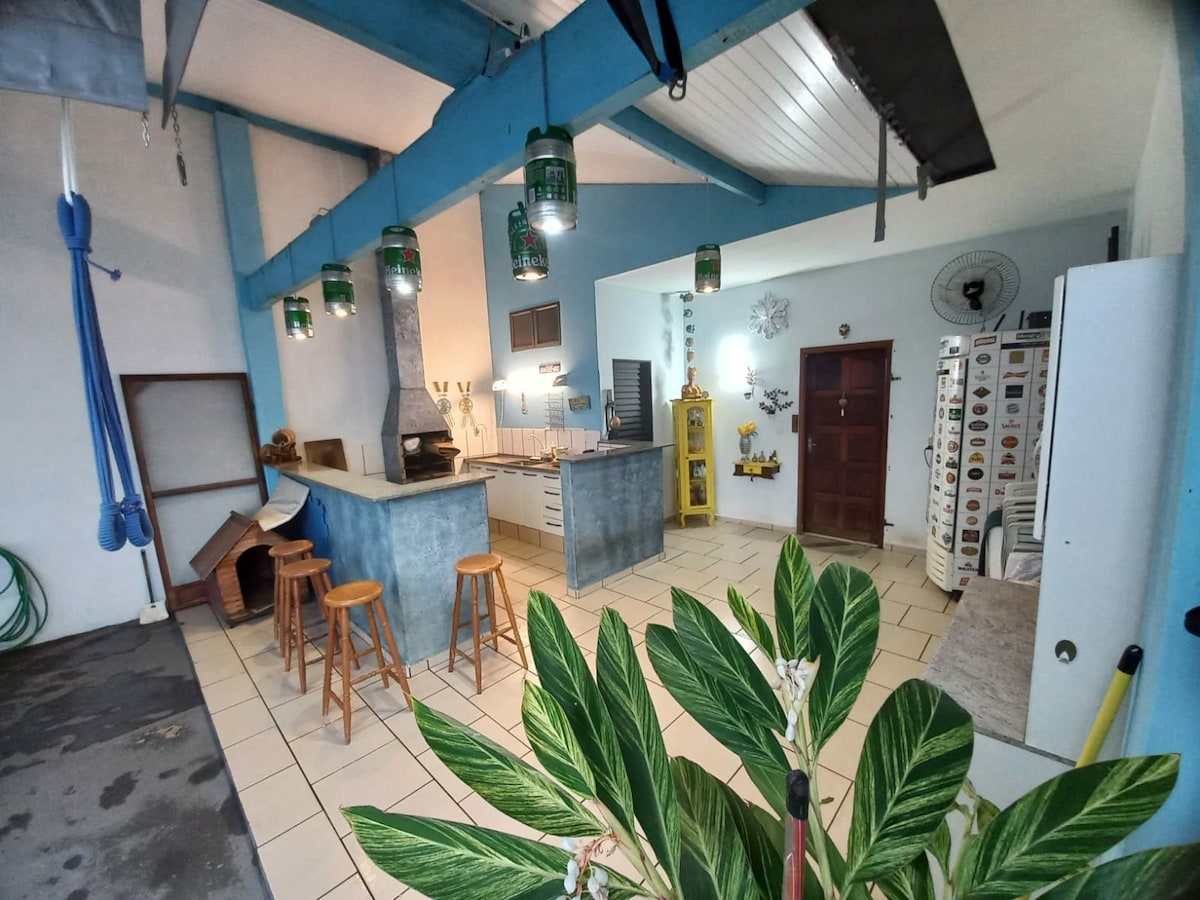 A spacious kitchen area features an island with bar stools, complemented by blue beams overhead. Green lantern-style lights hang above the counters, while a fireplace is positioned against a textured wall. The setting is brightened by natural light filtering through nearby windows.
