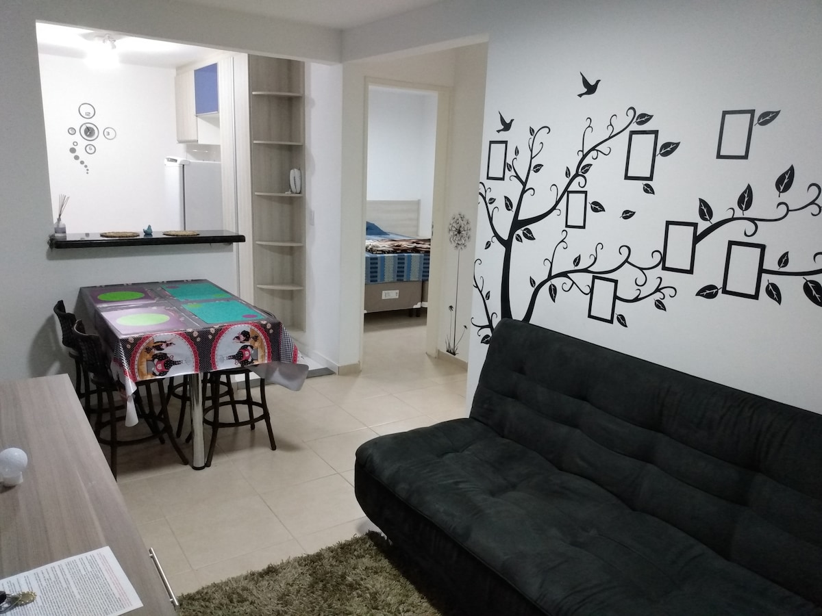 A spacious living area showcases a dark sofa and a dining table with colorful placemats. Wall art depicting birds and a tree enhances the ambiance. A light-colored kitchen can be seen in the background, along with hints of a bedroom through an open door.