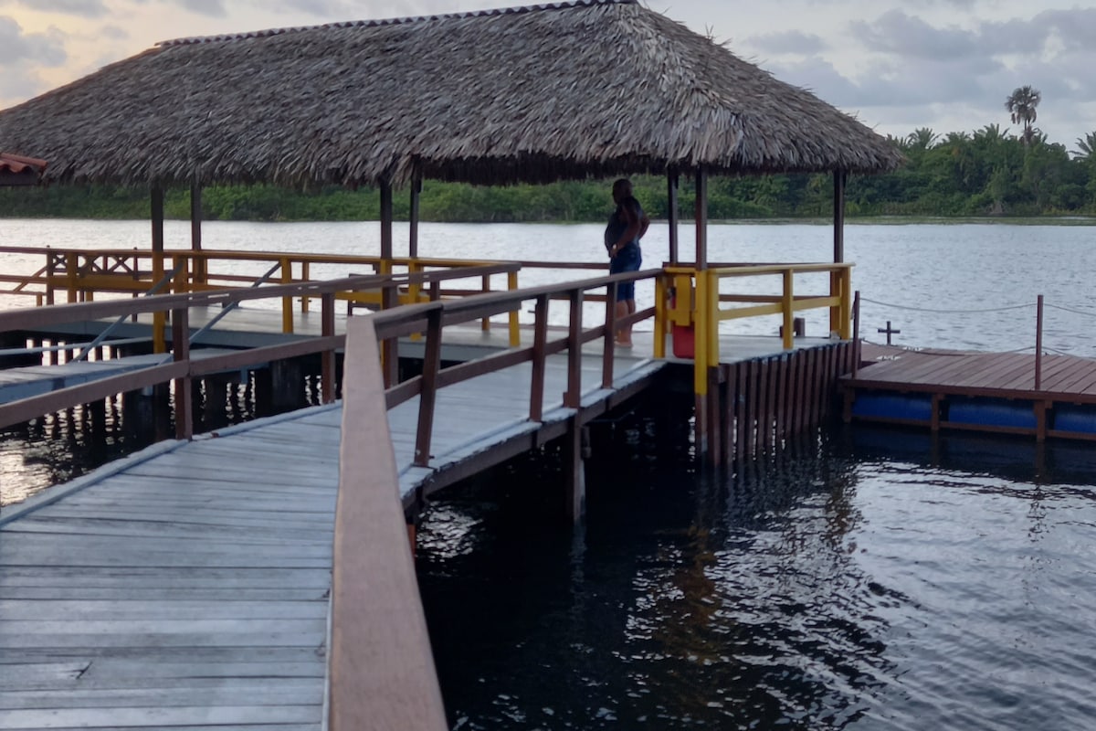 A covered pier extends over calm waters, featuring wooden walkways and seating areas. The thatched roof provides shade, and lush greenery can be seen in the background, enhancing the natural setting. The tranquil atmosphere invites guests to relax and enjoy the scenic views.