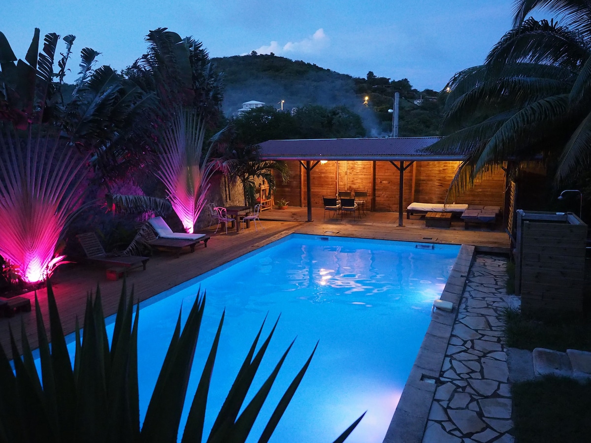 A well-lit salt-water pool reflects soft blue tones under the evening sky. Lush palm trees and vibrant pink lights frame the pool area, while shaded loungers provide a relaxing space for guests. A wooden deck surrounds the pool, enhancing its inviting ambiance.