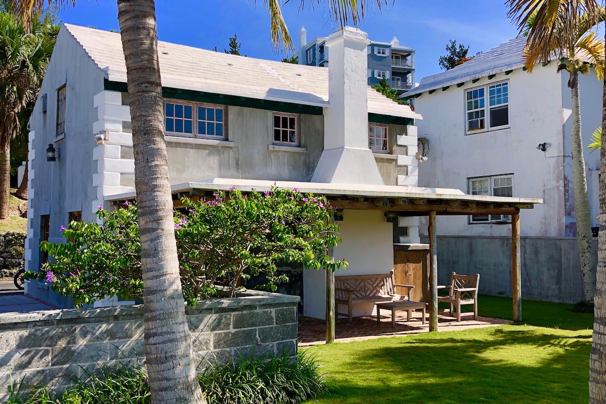 The exterior of Westport cottage is shown, featuring a covered verandah with seating. Lush green lawn and tropical palm trees are seen in the foreground, while the gray stone structure blends charmingly with surrounding buildings, showcasing a mix of historic and modern architectural elements.