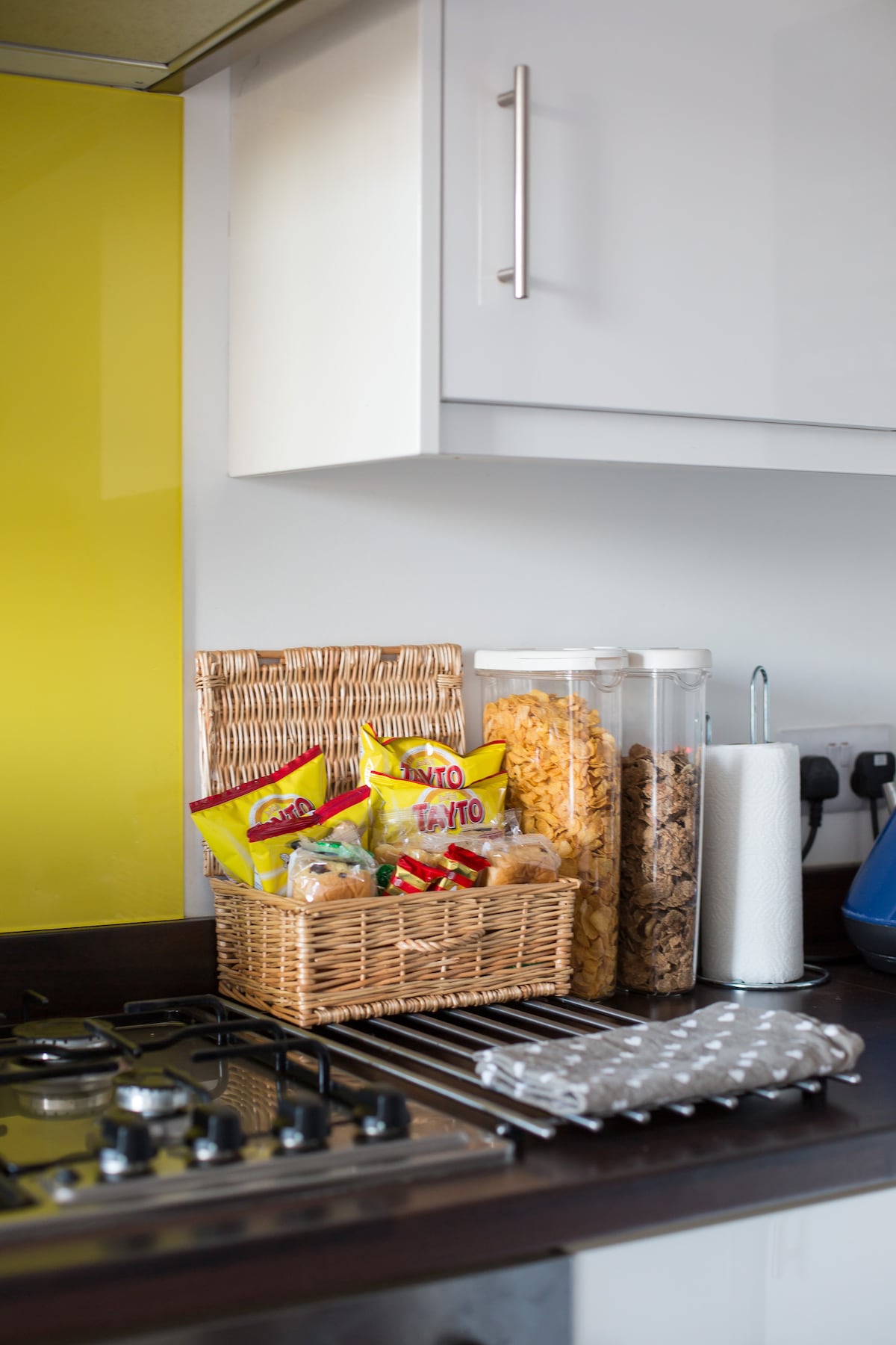 A modern kitchen area features a bright yellow wall and sleek white cabinetry. A woven basket holds snacks, while clear storage containers display cereals. A clean countertop is equipped with a paper towel holder and a patterned kitchen towel rests nearby.