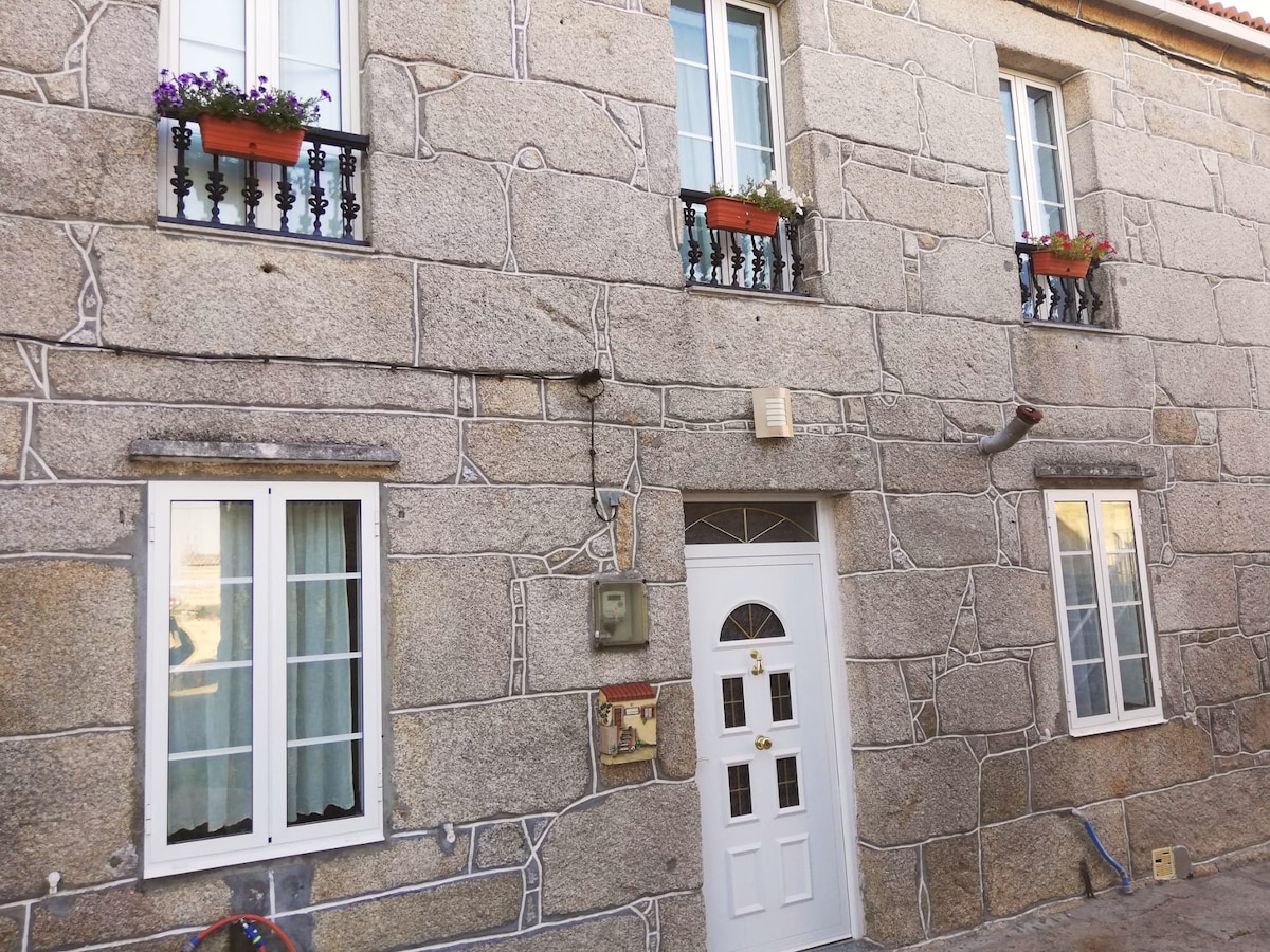 The exterior of a stone building is displayed, featuring a textured facade with prominent stonework. Four white windows are framed with black metal balconies adorned with flower boxes. A white door with a window design is positioned at the center. Soft natural light is evident.
