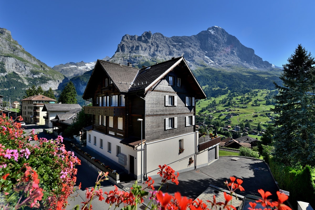 A charming wooden structure is set against a backdrop of majestic mountains, with lush greenery in the foreground. Bright flowers are visible in the foreground, complementing the building's inviting facade. The clear blue sky creates a tranquil atmosphere, enhancing the scenic view.