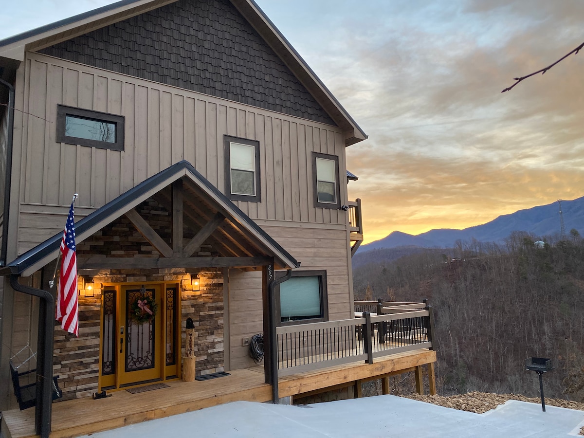 The cabin exterior features a welcoming entrance with a wooden deck and stone accents. An American flag is displayed beside the doorway. The background showcases distant mountains under a colorful sunset sky, highlighting the serene mountain setting.