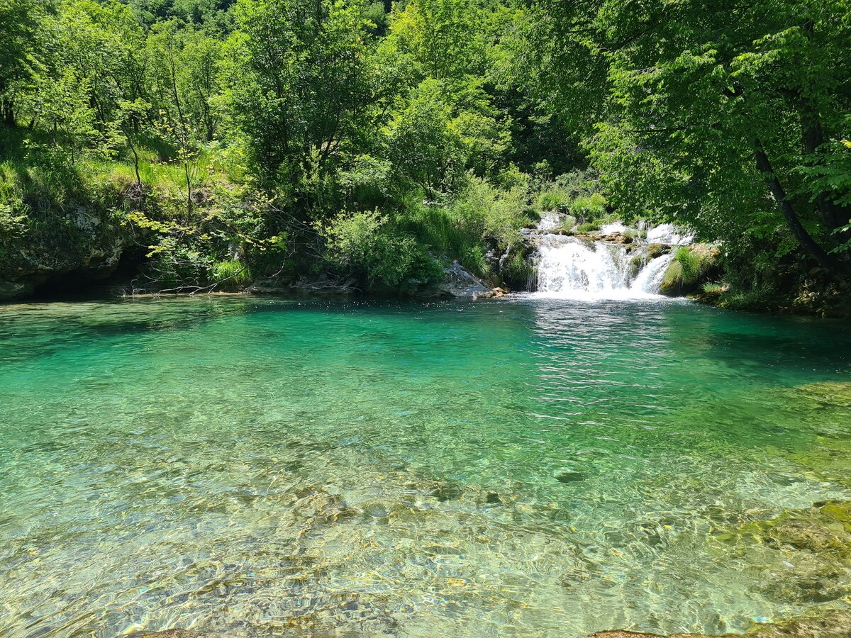 A clear, turquoise body of water is surrounded by lush greenery and trees. Small waterfalls can be seen cascading in the background, creating a serene natural setting. The water is shallow and reveals smooth stones at the bottom.