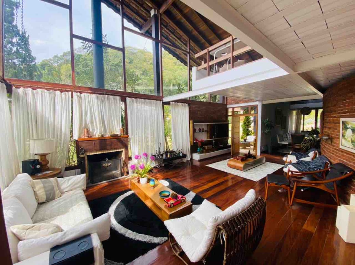 A spacious living area is filled with natural light, featuring large windows and wooden beams. Two comfortable sofas face a central coffee table adorned with decorative items. A cozy fireplace adds warmth, while lush greenery is visible through the glass doors.