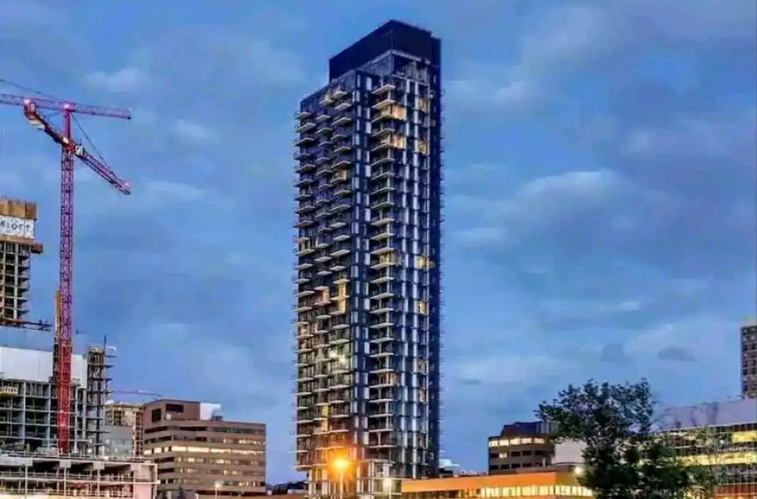 A modern high-rise building is depicted against a twilight sky. Its glass façade reflects the evening light, showcasing several floors of illuminated windows. Surrounding urban developments are visible, alongside construction equipment, suggesting ongoing growth in the area.