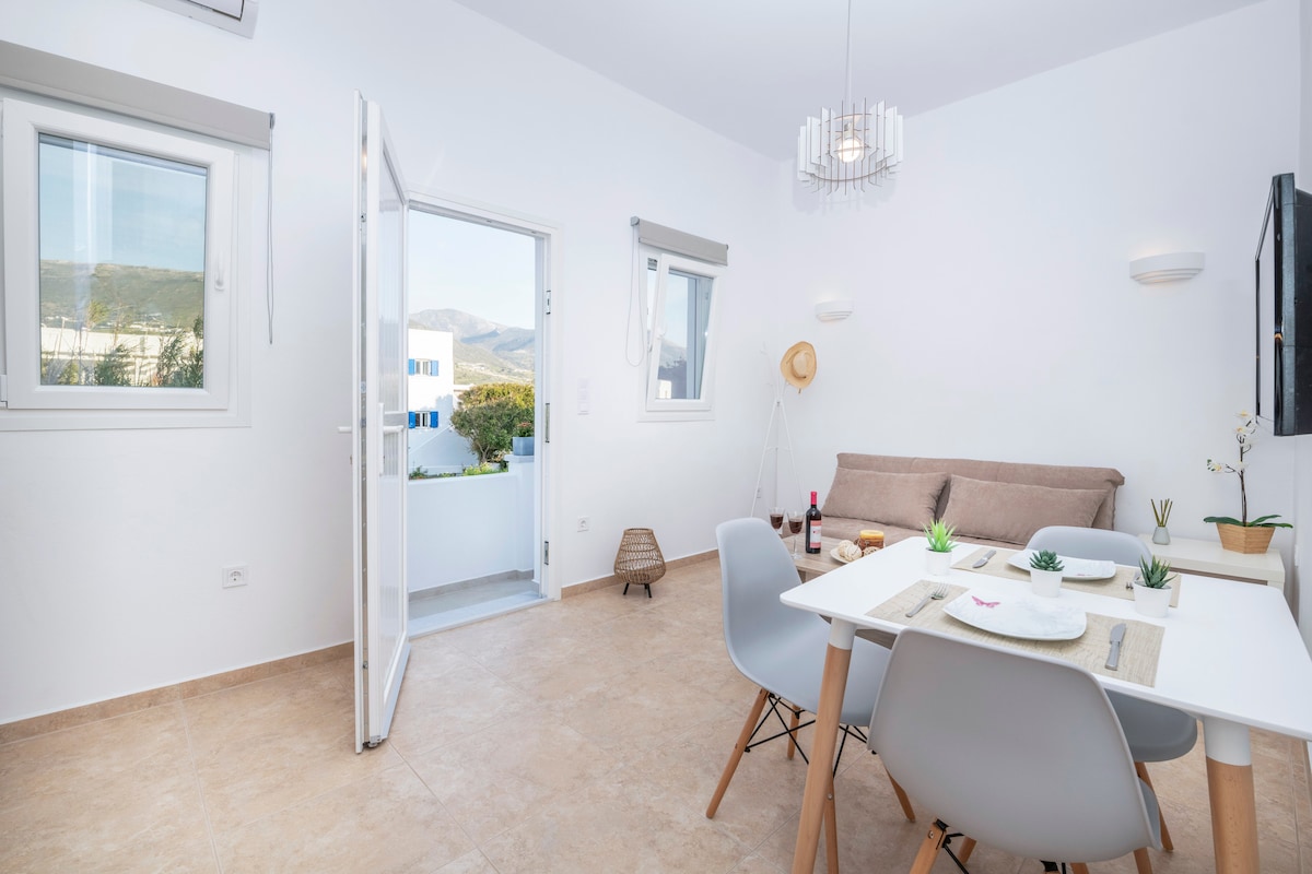 A modern studio space is illuminated by natural light from large windows. A dining table with four chairs sits beside a stylish sofa, while plants add a touch of greenery. The open door leads to a small private balcony, showcasing views of the surroundings.