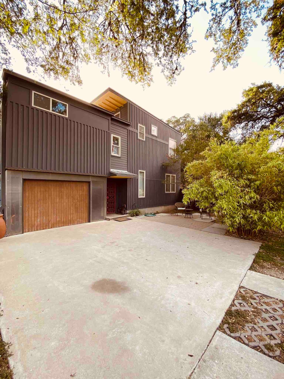 The exterior of the art studio features a modern two-story structure with vertical dark siding. A wooden garage door is visible, and a concrete driveway leads up to the entrance. Lush greenery is present, including a bamboo plant, providing a natural element to the scene.