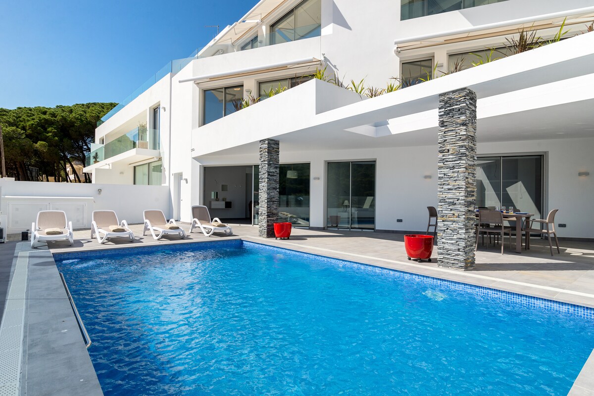 A private pool is surrounded by a spacious terrace, featuring several sun loungers and a dining area. The modern architecture is highlighted by stone columns and large glass doors that offer views of the pool area and abundant natural light.