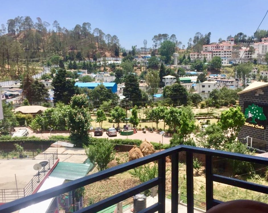 A scenic view from the balcony captures lush greenery and distant town structures, with trees dotting the landscape. The area below features various buildings, gardens, and a pathway, all nestled among the hills, offering a peaceful ambiance complemented by the surrounding natural beauty.