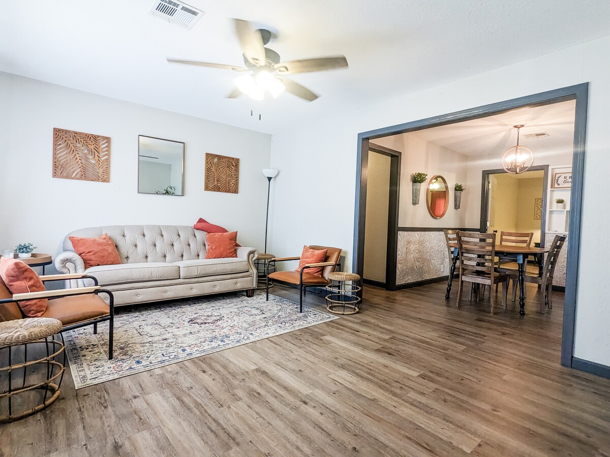 A spacious living area features a light-colored sofa adorned with orange cushions and a decorative rug. Two modern chairs complement the seating arrangement. A ceiling fan provides airflow, while mirrors and wall art enhance the ambiance. An adjoining dining area can be seen through the opening.