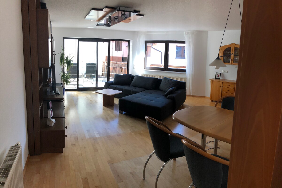 An open living and dining area features a spacious dark sectional sofa and a wooden dining table with four chairs. Large glass doors lead to a private balcony, allowing natural light to fill the space. A light-colored wood floor complements the room's modern design.