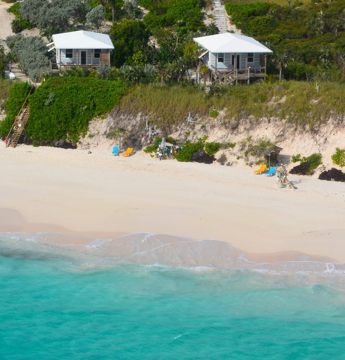 Two beachfront villas are nestled on a secluded pink sand beach, surrounded by lush greenery. Calm turquoise waters lap gently at the shore, while beach chairs and umbrellas are arranged invitingly on the sand, creating a tranquil retreat.