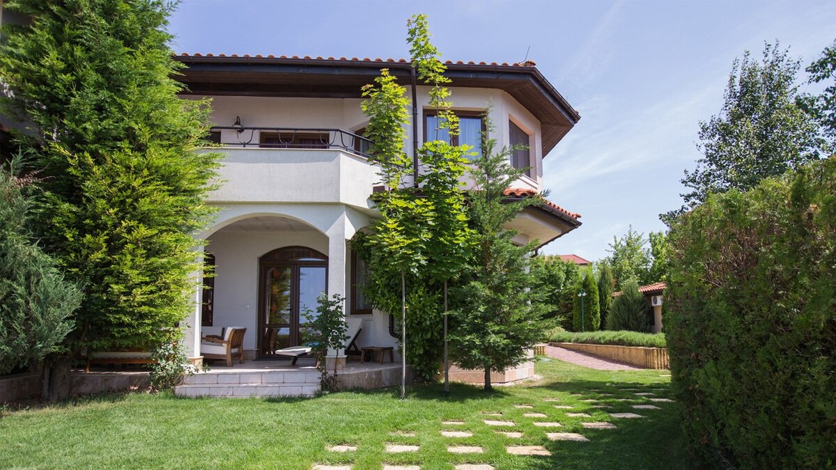 The two-story villa features a charming exterior with a combination of stone and stucco walls. Lush greenery surrounds the property, including manicured hedges and trees. A covered terrace with seating is visible, providing a relaxing spot to enjoy the garden view.