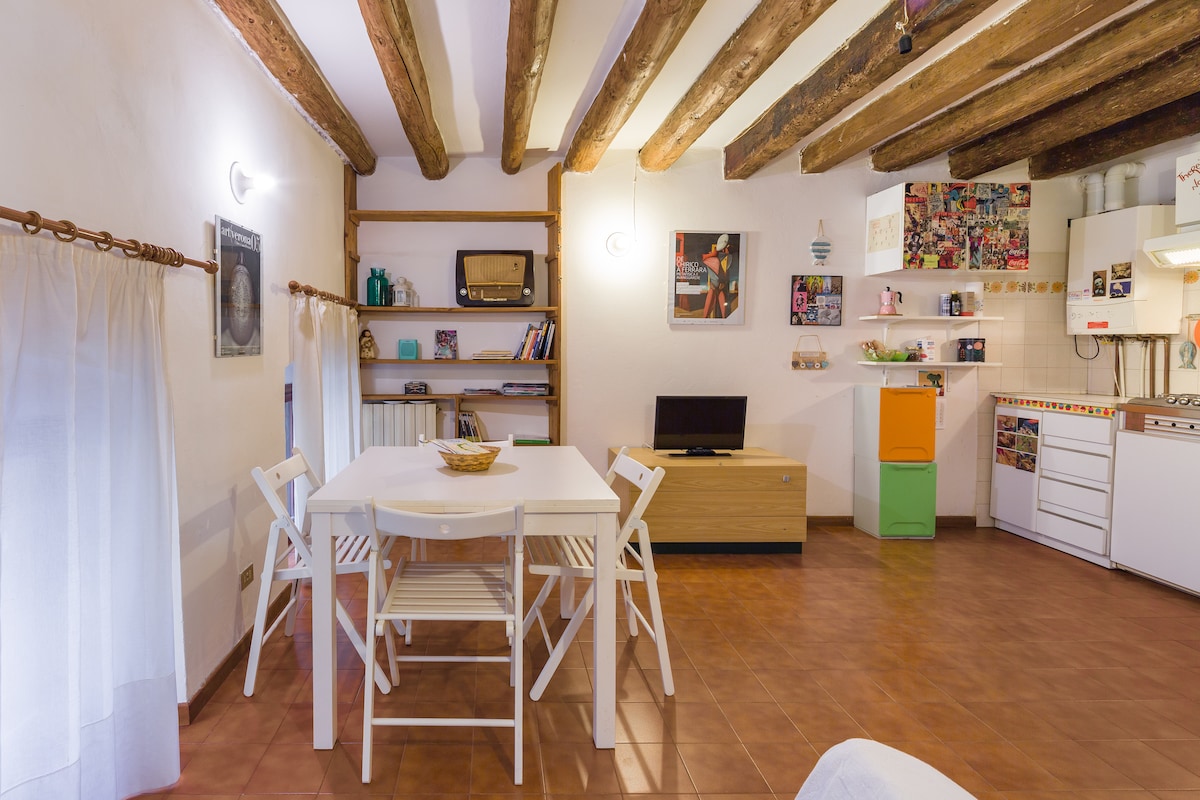 A cozy living area features a wooden dining table with white chairs, complemented by exposed wooden beams overhead. Shelving units display books and decorative items. A small television is positioned on a light-colored cabinet, with an equipped kitchen area visible in the background.