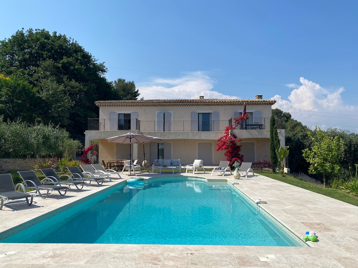 A Mediterranean villa is pictured, framed by lush greenery. A spacious salt-water pool, surrounded by lounge chairs, reflects the clear blue sky. A large umbrella provides shade near outdoor seating. Vibrant flower accents add color against the neutral facade of the home.