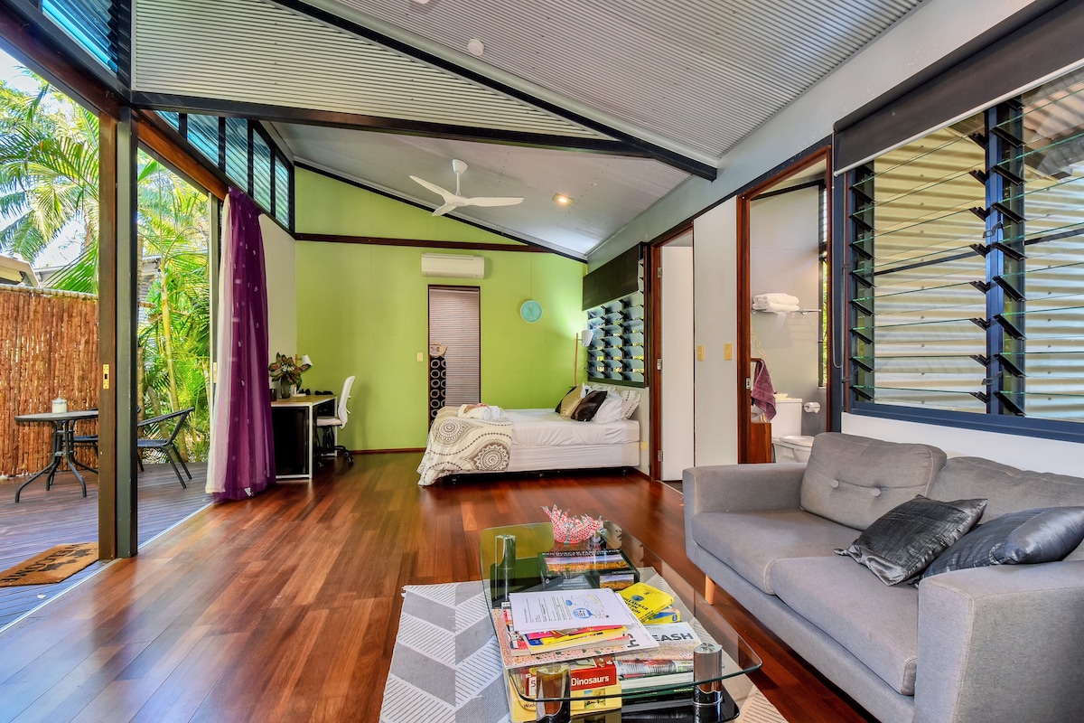 An open-plan living area features a spacious bed with light bedding, a cozy sofa, and a glass coffee table adorned with magazines. Large windows allow natural light to fill the space, showcasing wooden flooring and a vibrant green accent wall. A small desk is positioned nearby.