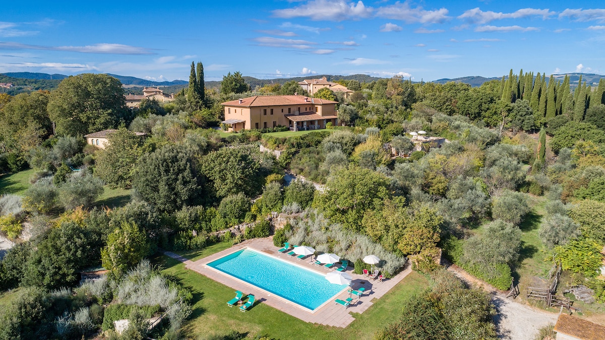 An aerial view showcases the villa nestled among lush greenery, surrounded by trees and shrubs. The rectangular swimming pool is prominently displayed with lounge chairs nearby, creating a serene outdoor area. The rolling hills of the Tuscan countryside provide a picturesque backdrop.
