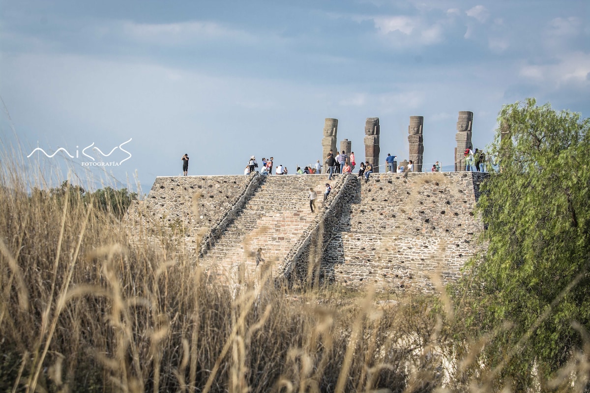 The archaeological site is depicted with several stone structures and towering sculptures visible. Visitors can be seen exploring the area, while surrounding tall grass and trees frame the location. The sky features soft clouds, contributing to the historical ambiance of the site.