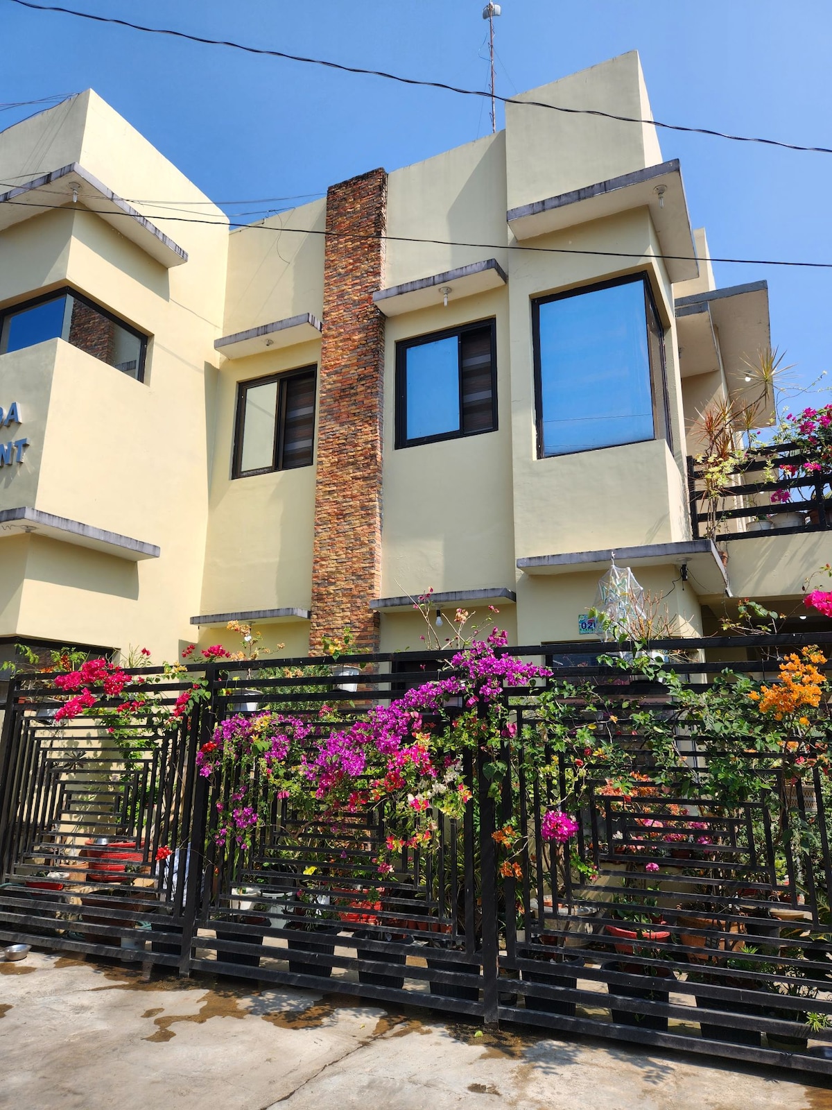 The exterior of the building is highlighted by a modern design with large windows. Colorful flowering plants cascade over the black wrought iron fence, adding vibrancy to the entrance. The sunny, clear blue sky complements the overall inviting appearance of the property.