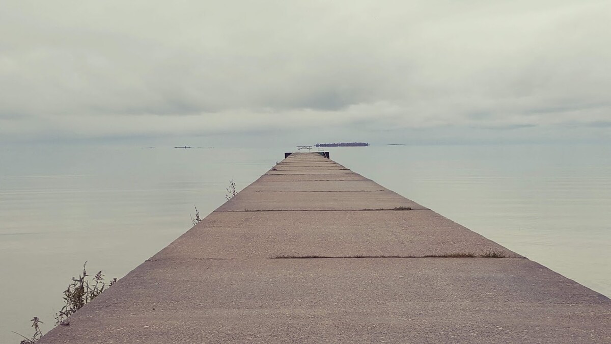 A long, concrete pier extends into calm waters, leading towards a distant island. Gray overcast skies suggest a tranquil atmosphere. The pier is bordered by sparse vegetation, and the smooth surface reflects the muted tones of the surroundings.