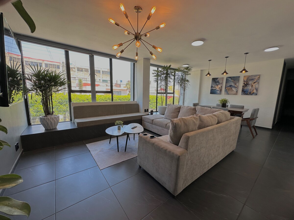 The spacious living area features large windows that allow natural light to fill the room, adorned with potted plants. A comfortable L-shaped sofa is positioned around a modern coffee table, alongside a dining area with overhead lighting and artwork on the walls.