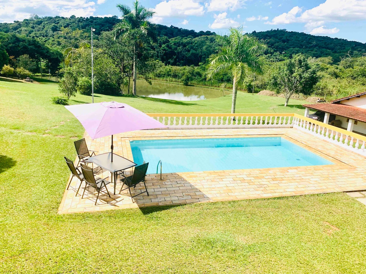 A swimming pool is positioned on a stone patio surrounded by a lush green landscape. A purple umbrella provides shade over a table with four chairs, while rolling hills and a tranquil lake are visible in the background under a partly cloudy sky.