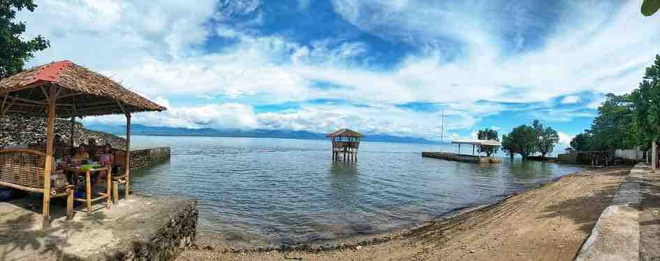 A panoramic view of the tranquil beachfront features a sandy shore leading to gentle waves lapping at the water's edge. A thatched-roof gazebo provides shaded seating, while a wooden platform extends over the water, surrounded by lush greenery and distant mountains beneath a blue sky.