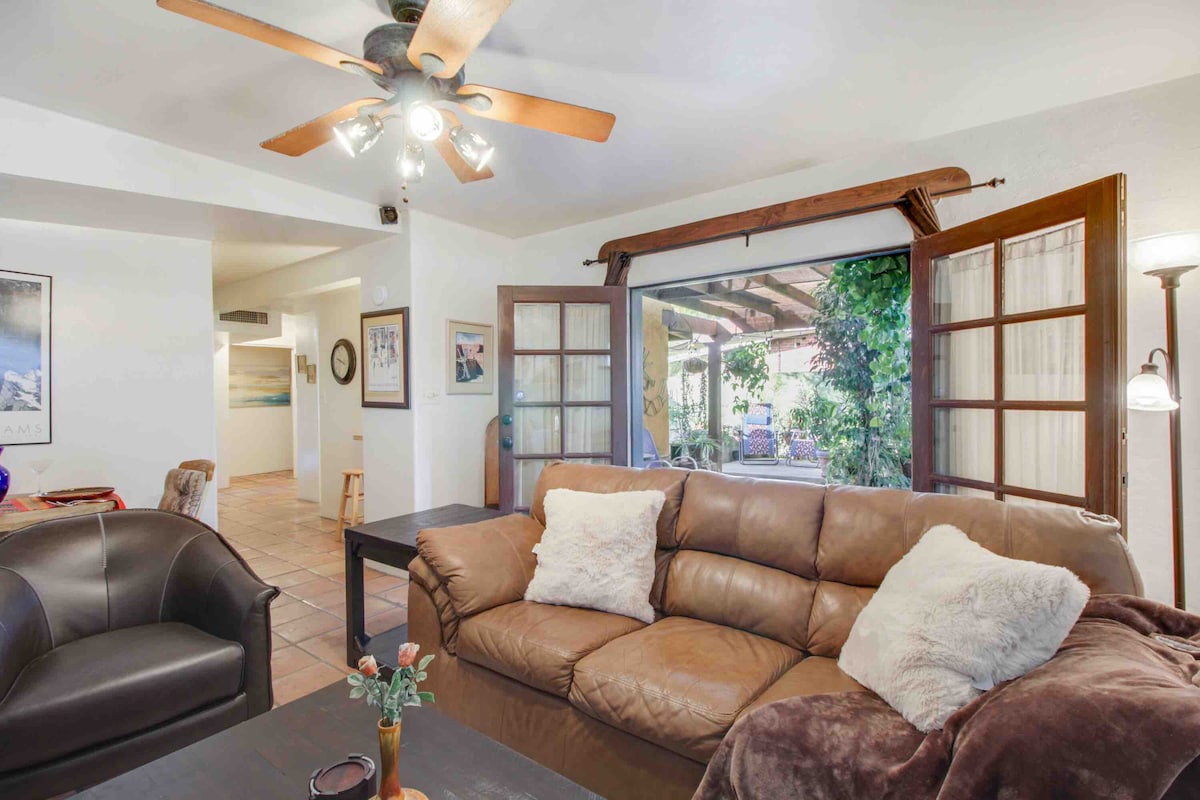 A comfortable living area is highlighted by a brown leather couch with decorative pillows. A wooden coffee table centers the space, while large windows allow natural light to illuminate the room. Greenery can be seen outside, creating a serene outdoor connection.