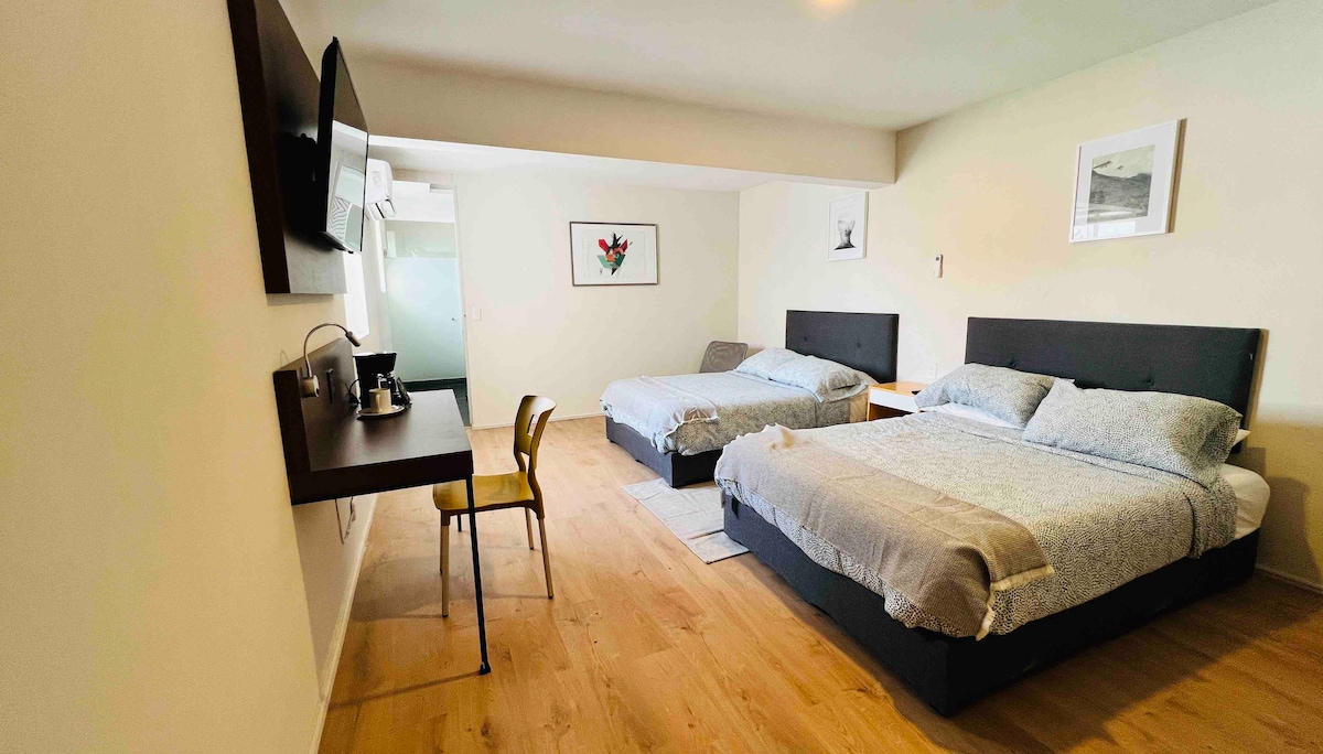 The room features two spacious beds with neutral bedding and a small desk with a chair for work or leisure. A television is mounted on the wall opposite the beds, enhancing convenience. Soft natural light fills the space, creating a comfortable atmosphere.